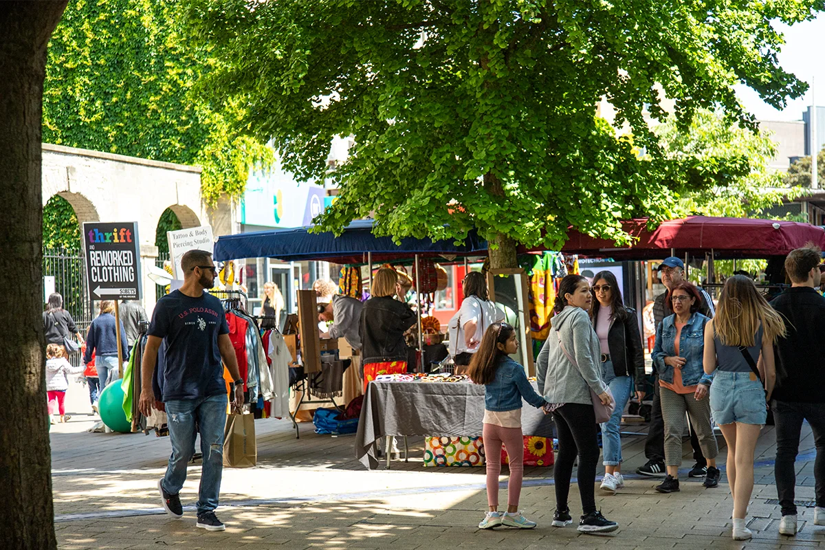 People walking around sunny Boardmead Sunday Market with large tree providing shade